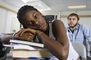 Closeup portrait of bored young African American student resting on stack of books in classroom