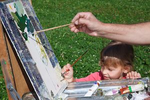 Easel artist in nature. Girl learns to paint with adult
