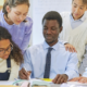 Diverse students gather around a male facilitator as he writes on papers at a table, collaborating on a project.