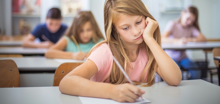 Young girl in a pink shirt writing at a desk in a classroom, with other students blurred in the background.