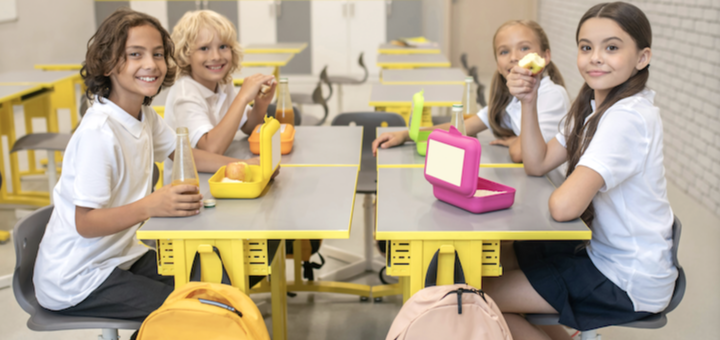 Group of four smiling students in a bright cafeteria enjoying lunch at yellow tables with lunchboxes and bottles nearby.